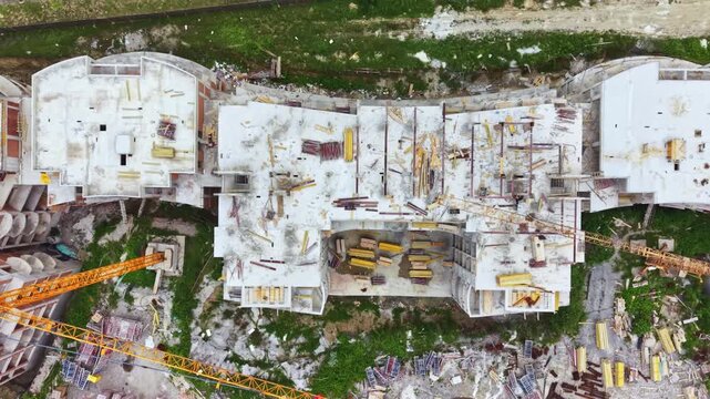 Aerial view construction site rooftop cranes showing concrete skeleton of multistory building under progress, heavy equipment parked on decks, scattered rubble