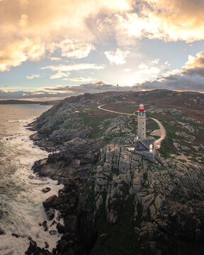 Aerial view of Faro de Punta Nariga lighthouse on a rugged rocky cliff with crashing waves and golden sunset light Malpica, A Coruna, Spain.