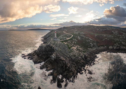 Aerial view of Faro de Punta Nariga on rocky cliffs with crashing waves and wind turbines under a cloudy sunset sky in Malpica, A Coruna, Spain.