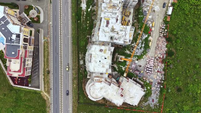 Aerial construction site beside road showing white concrete shells, excavator clearing rubble, curved foundation outlines, adjacent highway traffic, green field
