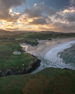 Aerial view of Ermita de Santa Comba on a green cliff overlooking the sandy beach and crashing waves during sunset in Covas, La Coruna, Spain.
