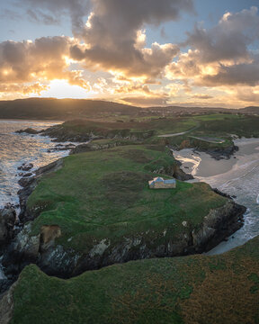 Aerial view of Ermita de Santa Comba on a green rocky peninsula surrounded by the ocean and coastal cliffs under a dramatic sunset sky in Covas, La Coruna, Spain.