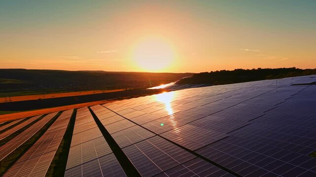 Sunlight shines on solar panels arranged in rows on a hillside. The sun sets in the background, casting a warm glow over the landscape.