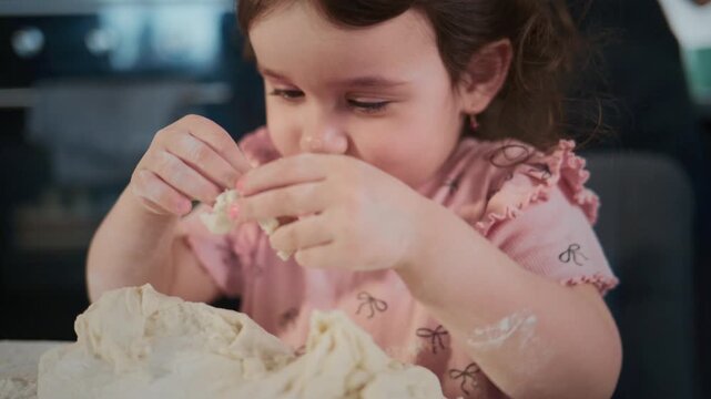 Young girl kneads dough with focused expression. She wears pink dress with bow pattern. Hands covered in flour show her playful effort. Scene captures joyful, domestic kitchen moment