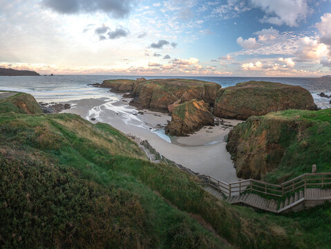 Aerial view of the Ermita de Santa Comba on a rocky islet surrounded by sandy beaches and rugged cliffs under a cloudy sky at sunset in Covas, La Coruna, Spain.