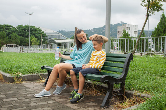 Pretty woman and child drinking pure water while resting on a bench in park after workout