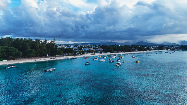 Aerial view of boats anchored in turquoise waters along the sandy shoreline of Trou-aux-Biches under a dramatic cloudy sky, Trou-aux-Biches, Pamplemousses District, Mauritius.