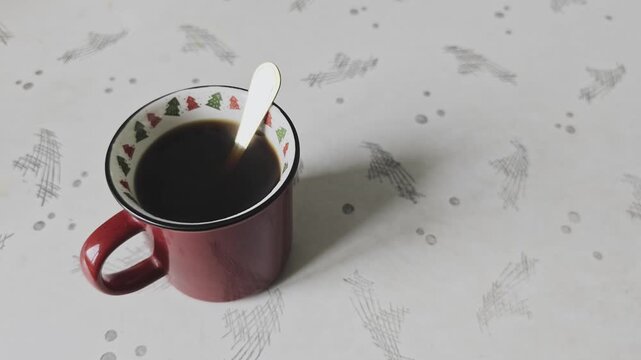 Red Christmas mug with hot black coffee and a spoon slowly rotating. Cozy holiday morning atmosphere with a warm beverage on a patterned table