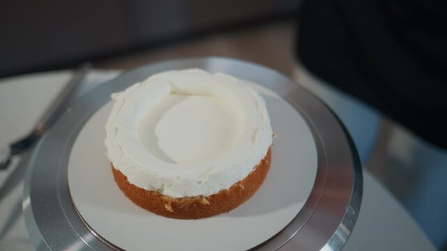 Baker applies smooth white frosting onto a cake. Hands rotate the cake stand for even coverage. The process shows careful, precise cake decorating. Ideal for culinary, baking