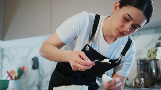 Young woman in black apron carefully slices layered cake. She leans in, focused, knife steady. Kitchen background with stainless steel tools. Suggests bakery, celebration, or dessert preparation
