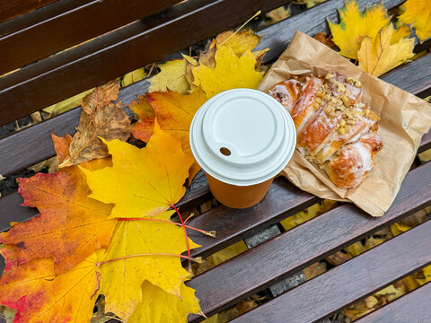 Paper coffee cup with hot drink and fresh croissant from bakery on bench covered with yellow fallen leaves from trees in autumn top view. Warming romantic coffee break in city park