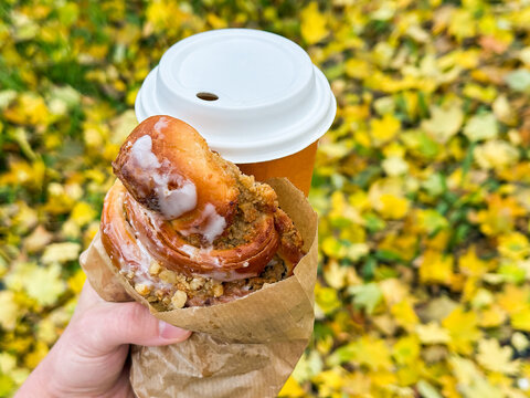 Woman hand with fresh pastry from bakery and hot coffee to go in paper cup with the background of ground fully covered with many fallen yellow leaves. Romantic walk in autumn park and coffee break