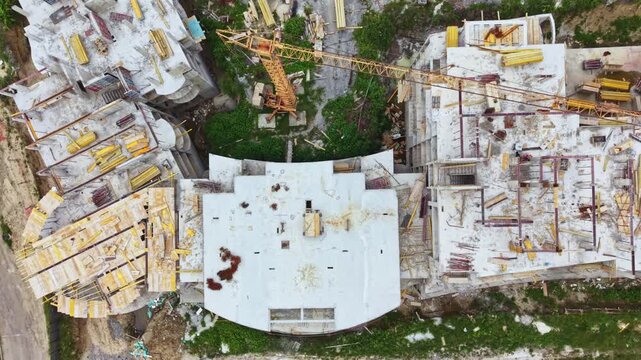 Aerial rooftop construction site showing weathered concrete rooftop, scattered materials and scaffolding, solitary worker inspecting surface, rusted rebar