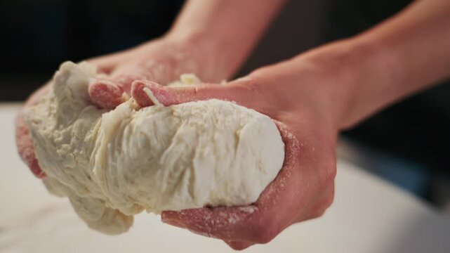 Hands knead soft dough on floured surface. Fingers press dough, showing texture and elasticity. Person focused, gentle touch suggests care and patience. Ideal for cooking, baking