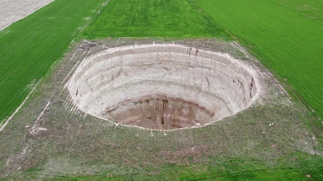 Drone footage showing a large sinkhole surrounded by green agricultural fields. Natural ground collapse, rural terrain, geological formation, and farmland landscape captured from above.