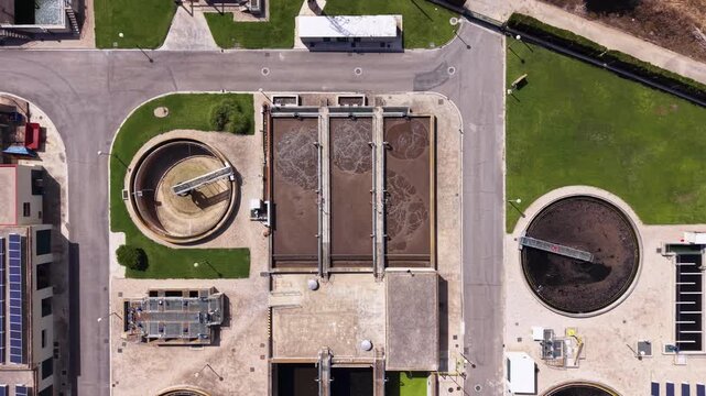 Top-down view of a wastewater treatment plant featuring circular clarifier tanks, aeration basins, solar panels, and green landscaping on a sunny day