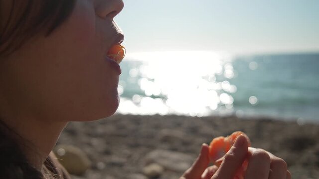 Tangerine beach eating woman enjoys a fresh fruit slice on the sunny shore during a relaxing summer vacation