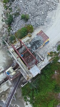 A truck is loading granite rocks from a quarry. Machines are operating to crush and transport materials. Workers are on site managing the processes during daylight.