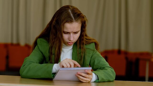 Focused student girl sits at desk and follows lines in notebook with finger, trying to memorize notes quickly for upcoming exam under pressure.