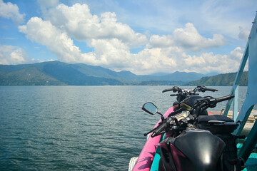 The handlebars and mirrors of motorcycles are in the foreground with a wide, calm lake and distant mountains stretching perfect for travel stories, transport, or themes focusing on exploration © DysOdyssey