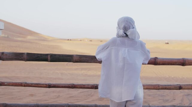 Woman farmer in white scarf observing desert landscape from camel farm wooden fence