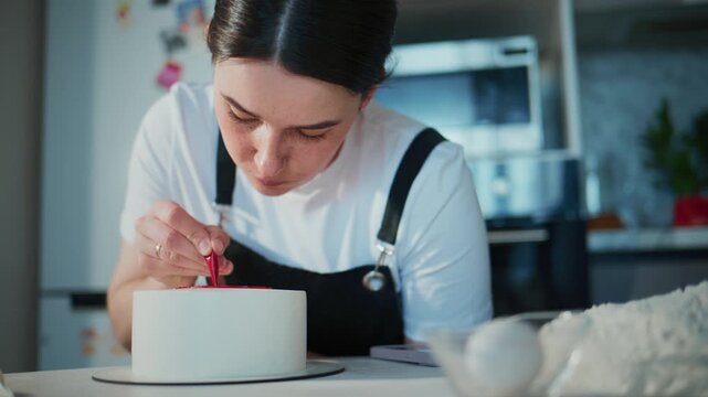 Focused baker decorates a white cake with red piping. She leans in, concentrating on precise details. Kitchen tools and flour surround her workspace. Ideal for recipes, baking tutorials
