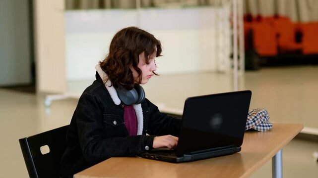 Teenage student with curly hair and wireless headphones around neck works on laptop in open study space, preparing project presentation and searching information online, concept productivity and self