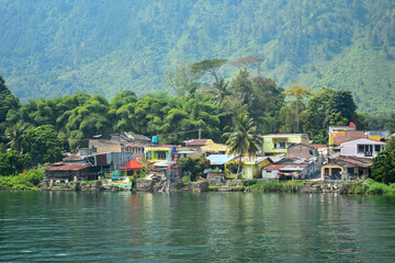 Obraz premium Colorful row of houses situated directly on the edge of a reflective lake, set against a backdrop of a tree-covered mountain, showcases lakeside human habitation perfect for geography, social studies