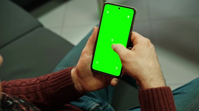 Hand of young man holding smartphone green screen in business office work. Mobile phone vertical mockup. Male person using phone with blank green background display. Chroma key smart phone. Close up.