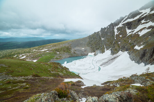 Scenic top view from green grassy slope to frozen lake among rocks and snows in mountain cirque against hills far away. Ice floes on turquoise water of alpine lake under cloudy sky in rainy weather.