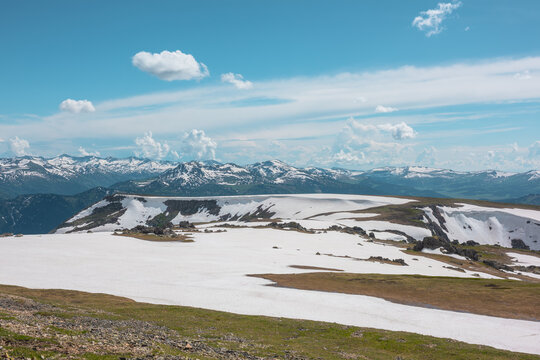 Scenic alpine landscape with sunlit snowy field on stony hill with view to big mountain range with forest and snow far away under cloudy sky. Stone outcrops among snows in sunlight in high mountains.