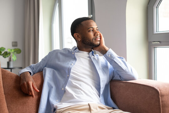 A young, mature African-American man sitting on the couch at home looking out the window daydreaming. Thoughtful handsome male relaxing in a minimalist living room