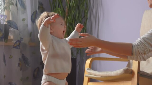 A baby takes first steps walking towards mother who has her arms wide open. Tender family moment of and joy