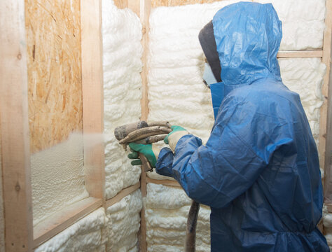 A construction worker in overalls applies insulation foam to a wooden wall of a house, house construction and insulation