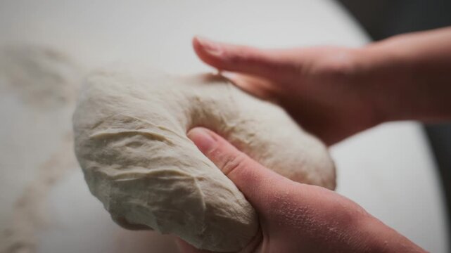 Hands gently knead fresh dough on a floured surface. Fingers press into soft, elastic dough. Texture shows rise and moisture of homemade bread. Close-up focuses on tactile process of dough preparation