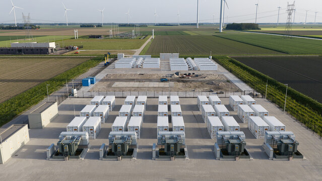 Aerial view of a large battery energy storage system facility with rows of white containers and wind turbines across flat agricultural fields in Dronten, Flevoland, Netherlands.