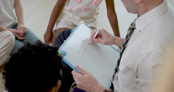 Close classroom circle view follows male adult teacher holding clipboard while young pupil group answers questions, checking first day routine, class rule, listening skill and back to school readiness
