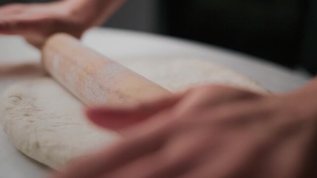 Person rolls dough with a wooden roller on a white surface. Hands apply gentle pressure as the roller flattens the dough. The focus stays on the motion and texture of the dough