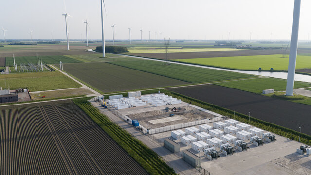 Aerial view of a battery energy storage system with white container units surrounded by agricultural fields and wind turbines in Dronten, Flevoland, Netherlands.