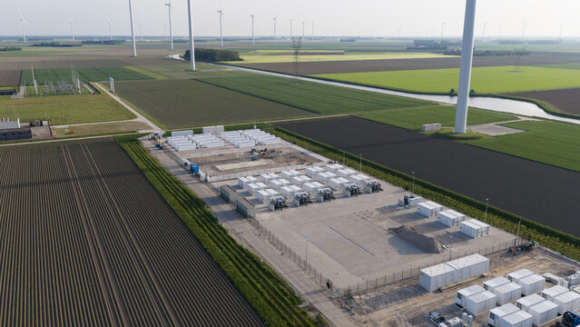 Aerial view of a battery energy storage facility with white containers and wind turbines across agricultural fields in Dronten, Flevoland, Netherlands.