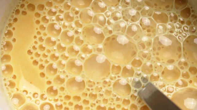Golden milk foam bubbles moving in circular motion inside mug. Stainless steel spoon rests still in the cup after stirring warm beverage made with organic turmeric powder