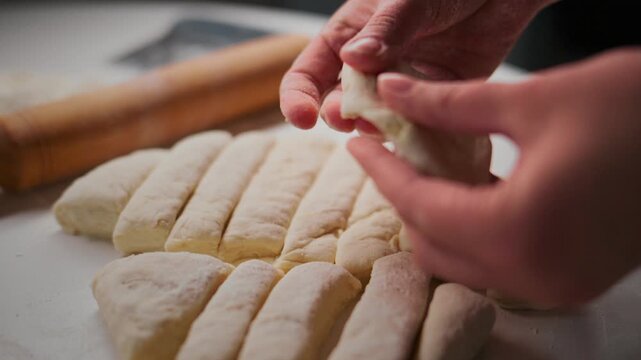 Close-up hands shape dough into logs on floured surface. Fingers press gently, forming rustic bread or pastry. Rolling pin rests in background, hinting at baking process. Ideal for recipes, food blogs