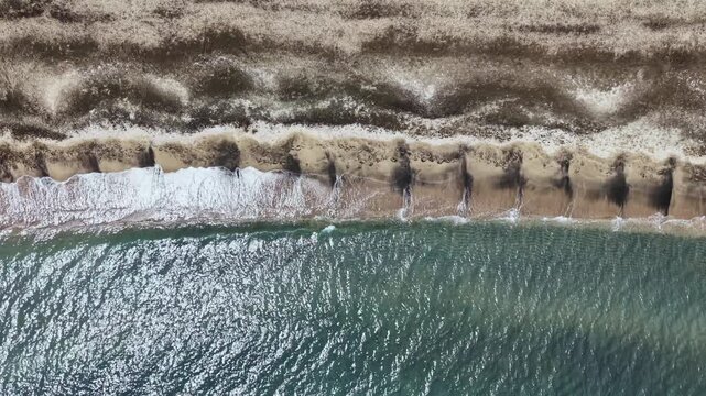 Aerial shoreline with glittering seawater and dunes, calm turquoise surf lapping narrow beach, rhythmic wave patterns, dark kelp lines, sandbars and erosion