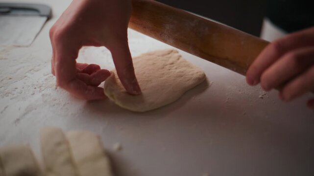 Person rolls dough with wooden roller on floured surface. Hands guide roller gently, ensuring even thickness. Dough stretches slightly under pressure, showing elasticity. Scene suggests home baking