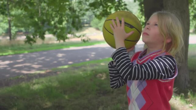 A beginner girl throws a basketball toward the hoop in a sunny park. She watches the ball flight and gets upset after missing