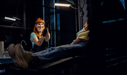 Fototapeta premium Female tattoo artist in a modern studio puts on gloves and prepares to make a tattoo on a female client.