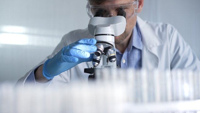Male scientist researcher wearing blue gloves, protective glasses is using microscope in modern laboratory, portrait view. Low key lighting photo. Science and Medicine concepts