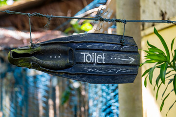 Creative toilet sign made from old diving flipper on tropical beach. Handmade rustic toilet navigation sign painted on a black rubber diving fin hanging outdoors in beach resort, Thailand © OlegD