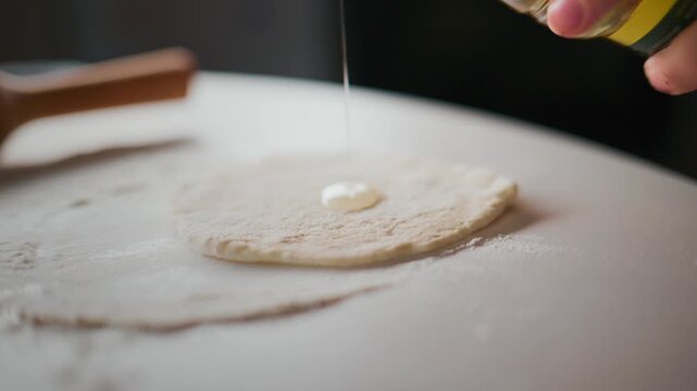 Hand sprinkles oil onto a flat dough circle. Dough rests on a floured white surface. Rolling pin lies nearby, unused. Scene shows calm, focused cooking preparation. Ideal for food blogs, recipe videos