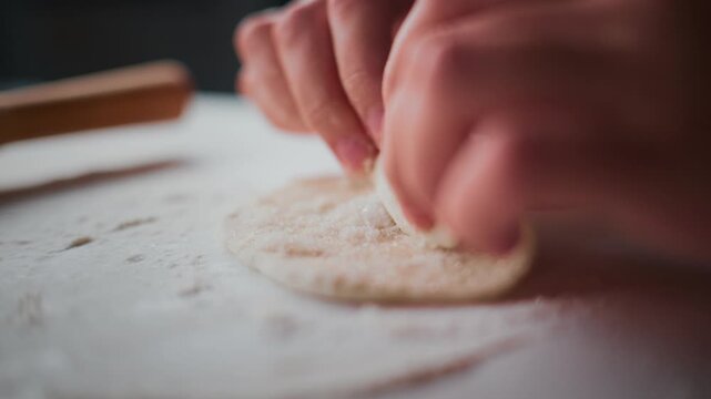 Close-up of hands shaping dough on flour-dusted surface. Fingers press gently, forming round base. Soft lighting highlights texture and motion. Perfect for food tutorials, baking content